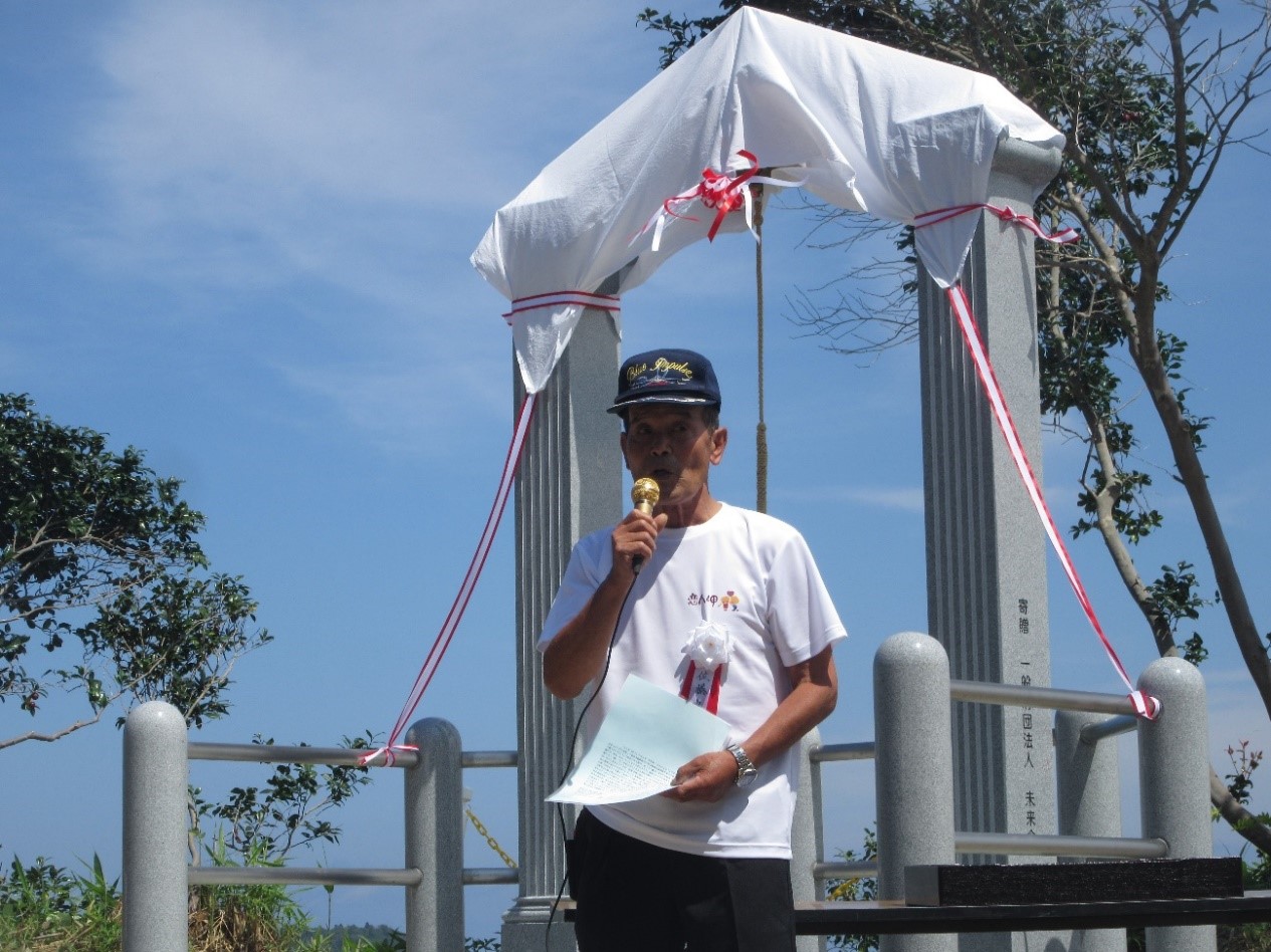 佐藤良夫さんが白い布で覆われた石碑の前に立ち、片手にマイクを持ち話している様子の写真