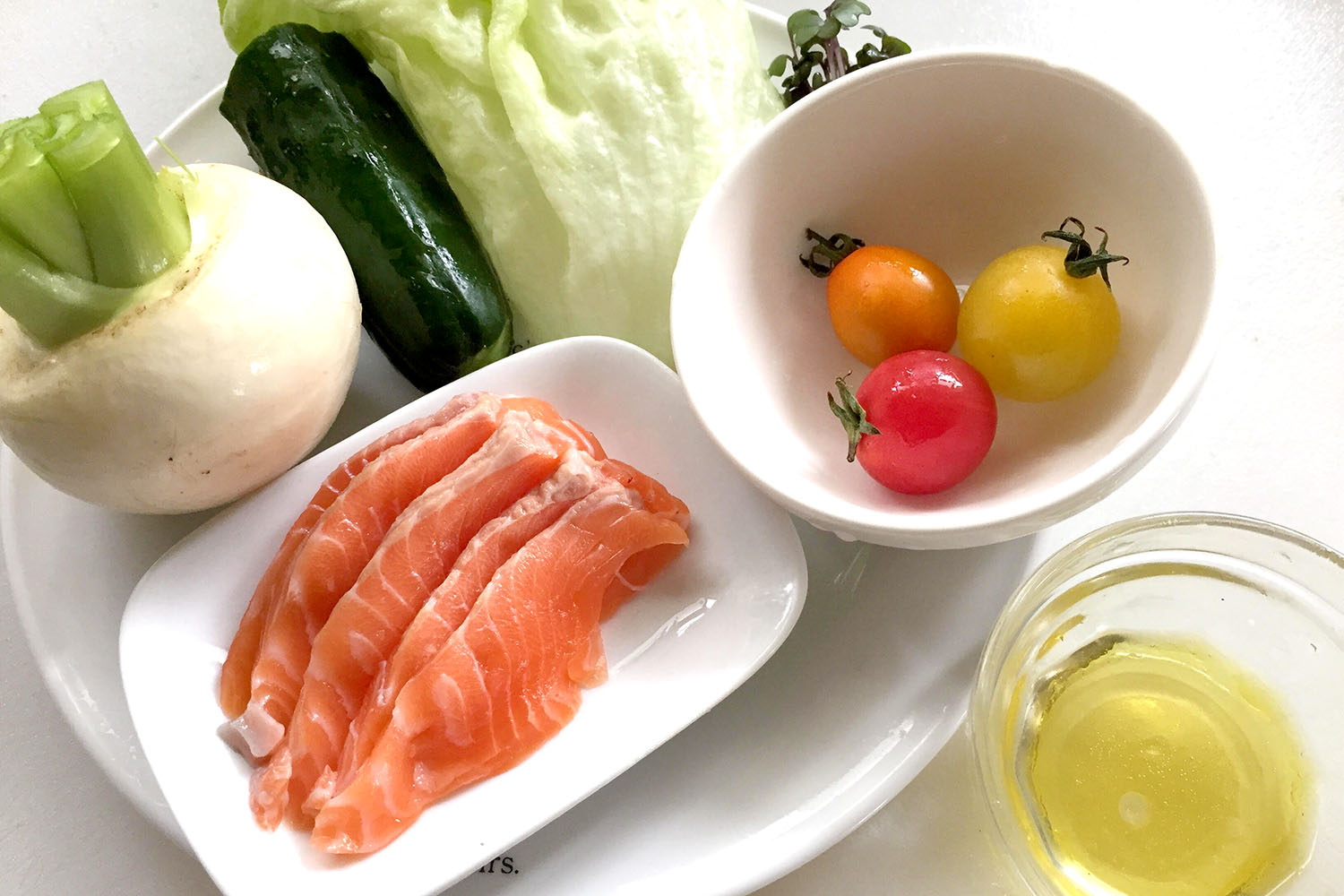白い皿に並べられた薄切りの鮭と、生野菜のカブ、キュウリ、レタス、3色のミニトマトと小さな器に入ったオリーブオイルが写っている食材の写真