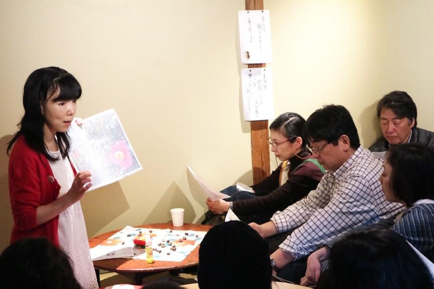 ちゃぶ台に椿の種が置かれた室内で、工藤さんが紙を見せながら復興のストーリーを伝えており、参加者たちが真剣に話を聞きながら資料に目を通しているお話会の写真