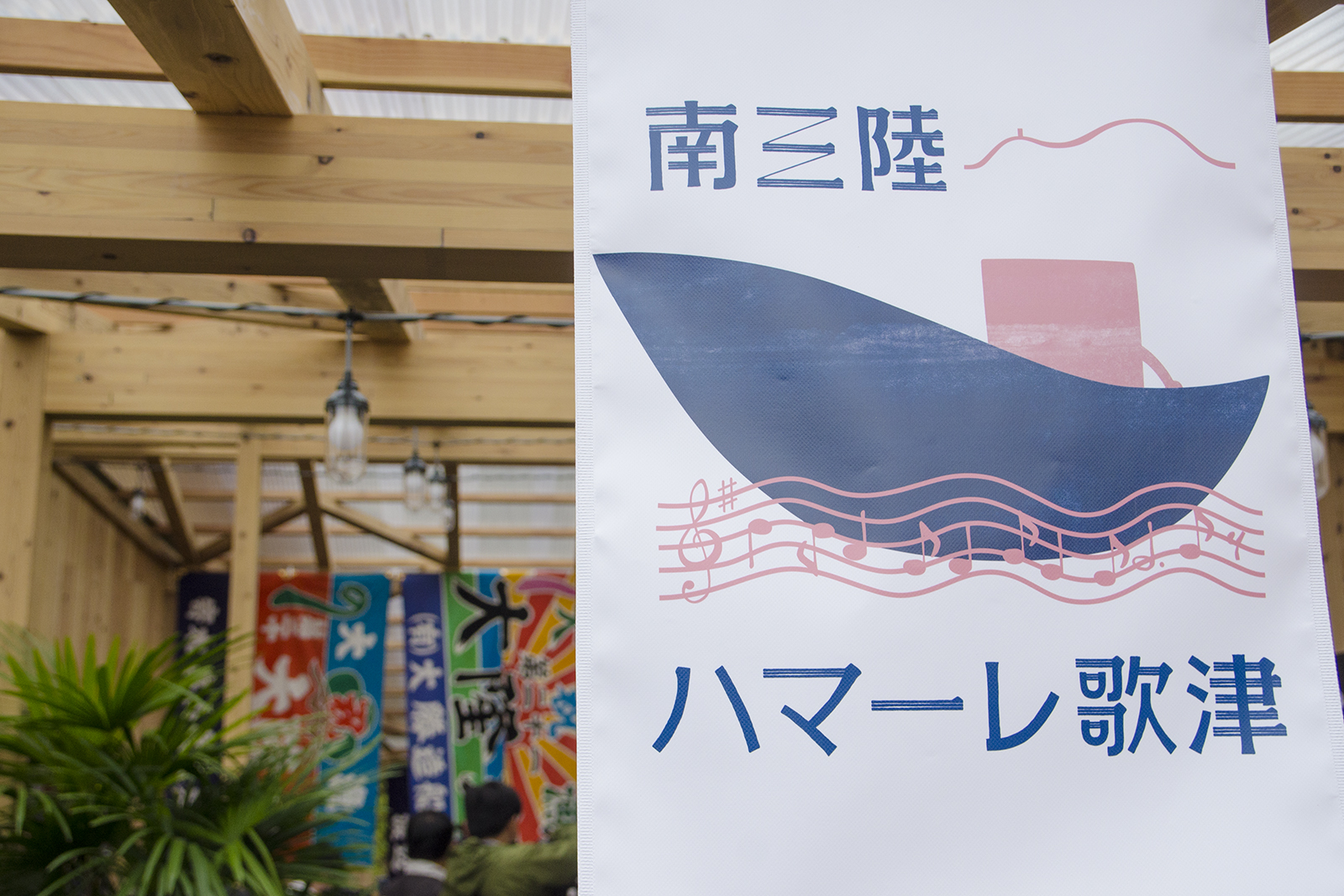 波型の楽譜の上に大きな船が浮かび、山並みのラインがデザインされ「南三陸 ハマーレ歌津」の文字が書かれた旗が商店街の軒下に飾られている写真