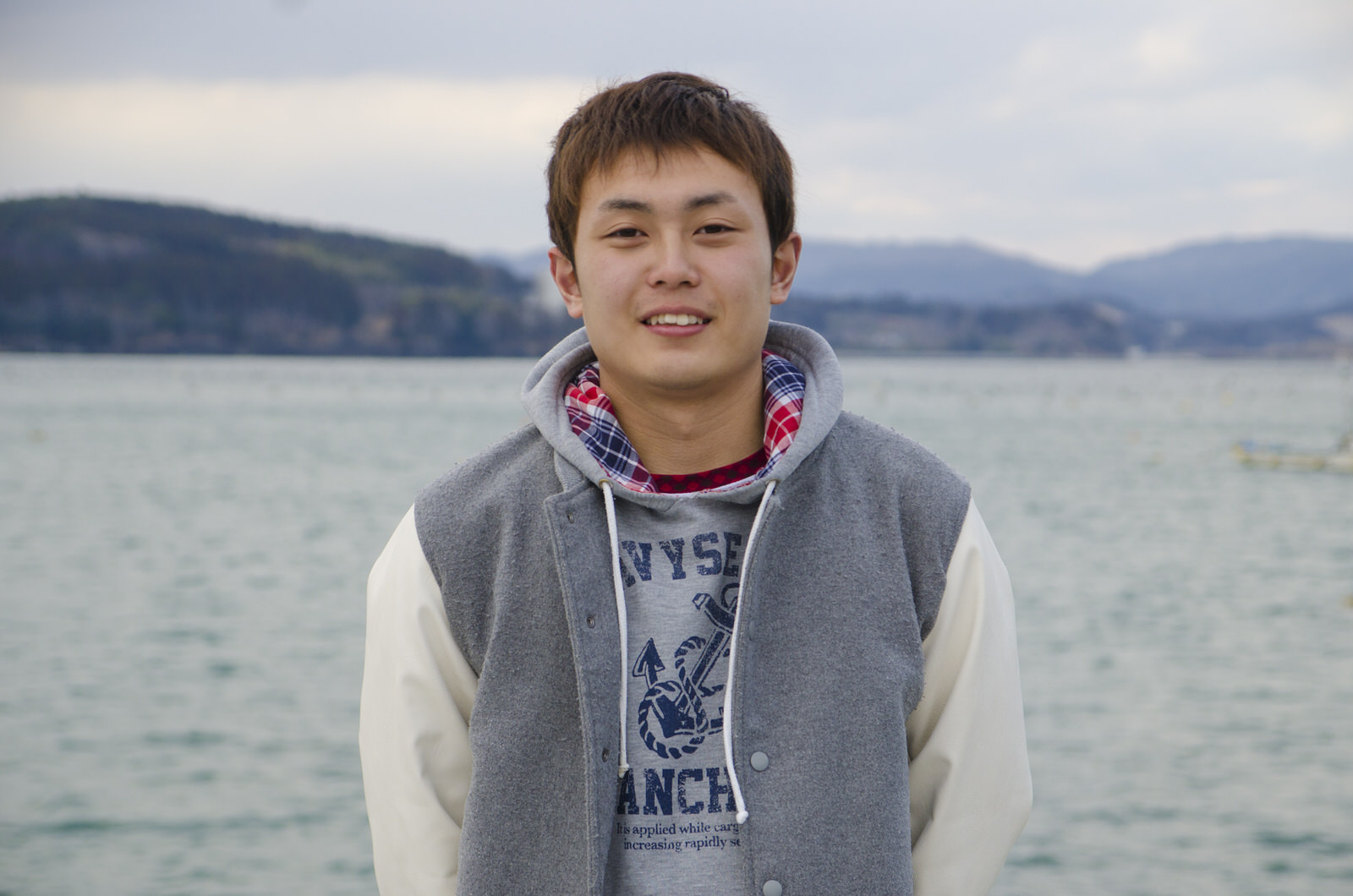 グレーと白の配色のジャンバーを着た三浦さんが海を背に笑顔で立っている写真