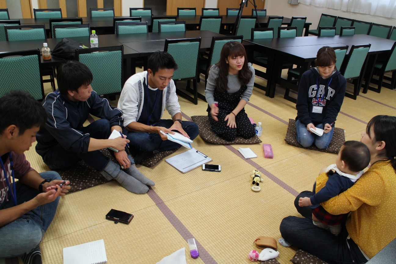 畳の部屋で、数人の人物が円形に座布団に座り、向かい合って話し合いをしている様子が写っている写真