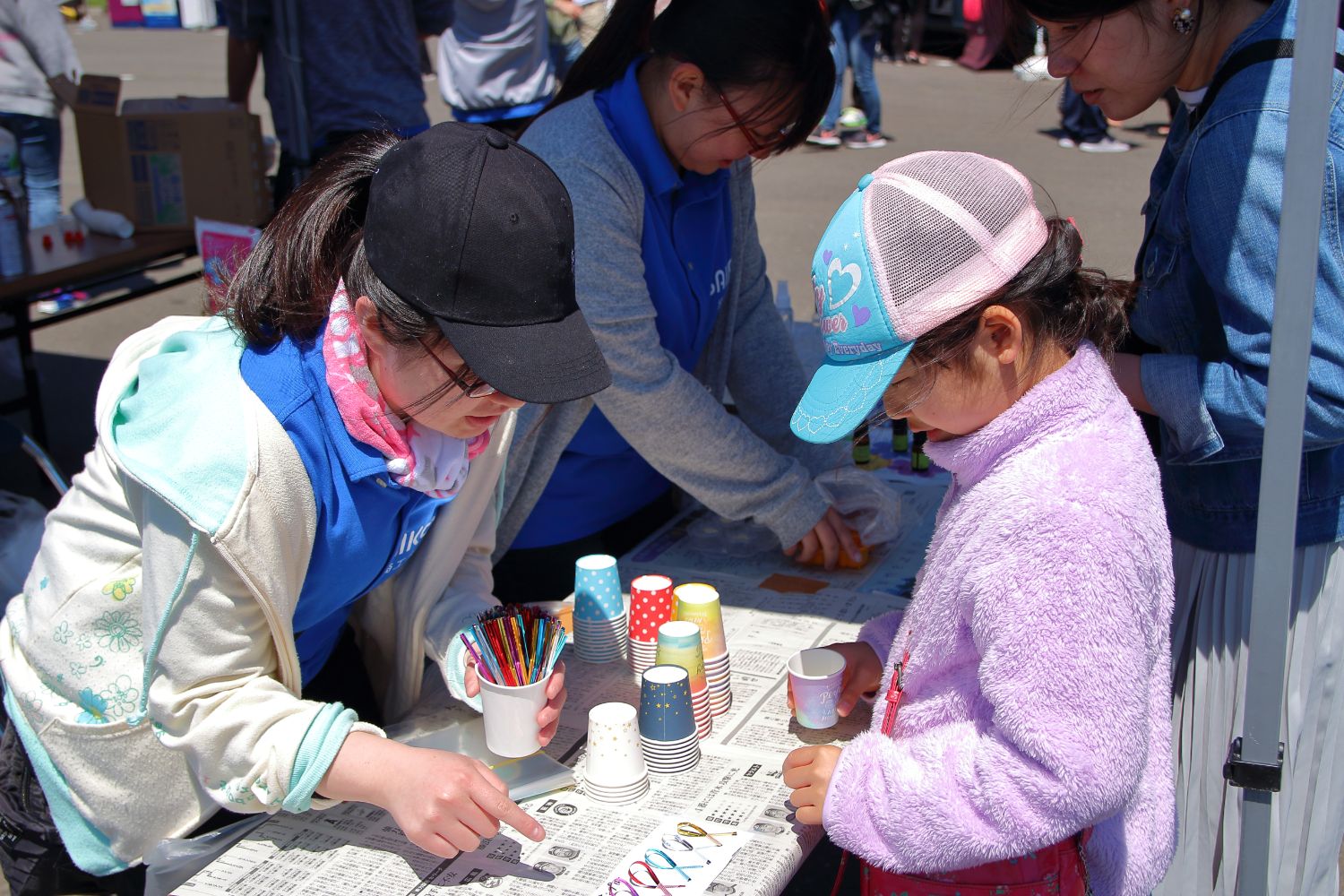 カラフルな紙コップなどが並べられたテーブルでに参加者の親子連れが立ち、向かい合って立つ女性スタッフに教わりながら女の子が色とりどりのリボンを選んでいる写真