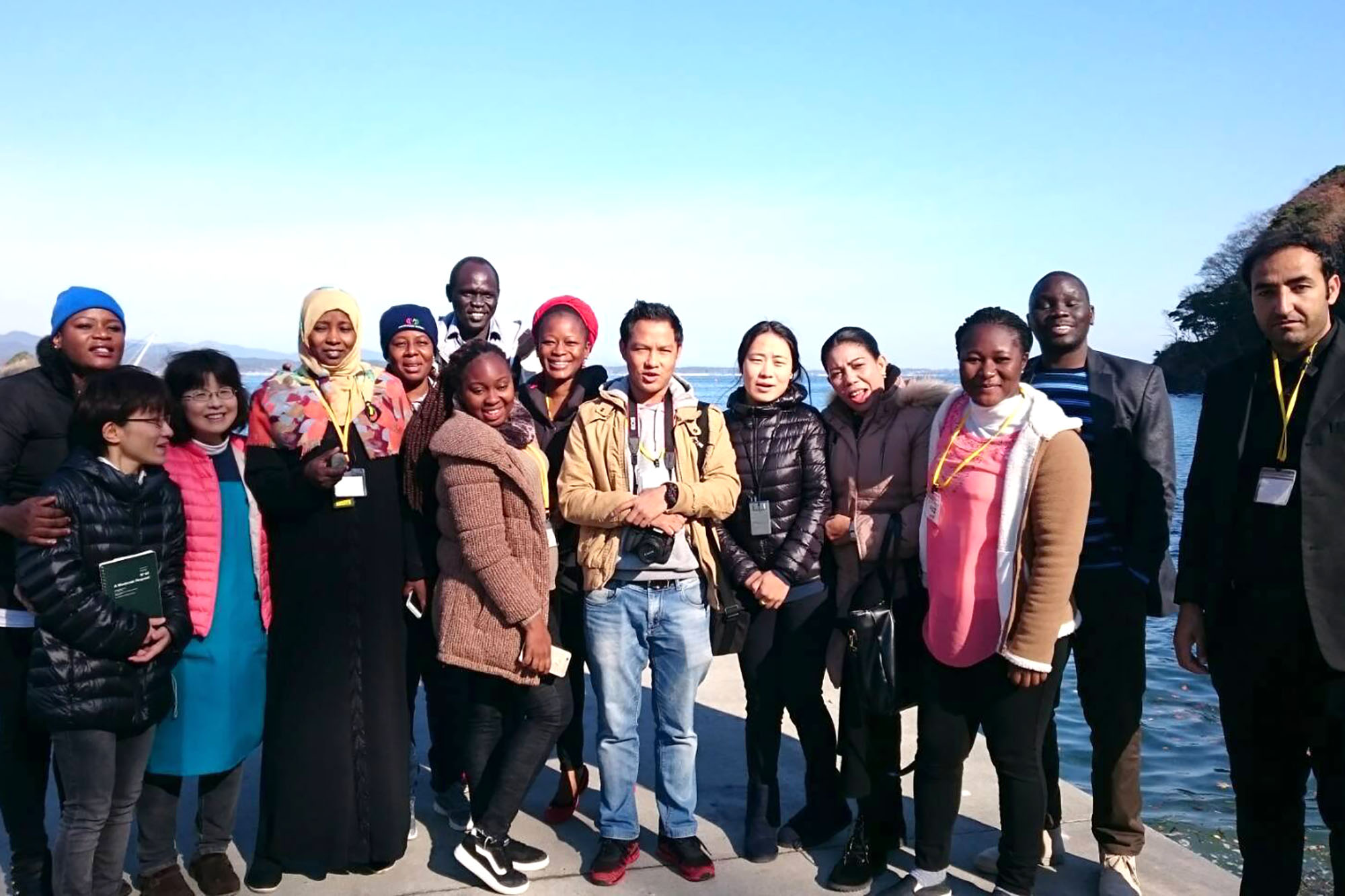 海を背景に、さまざまな国や地域から来たと見られる人々と阿部民子さんが笑顔で並んでいる集合写真