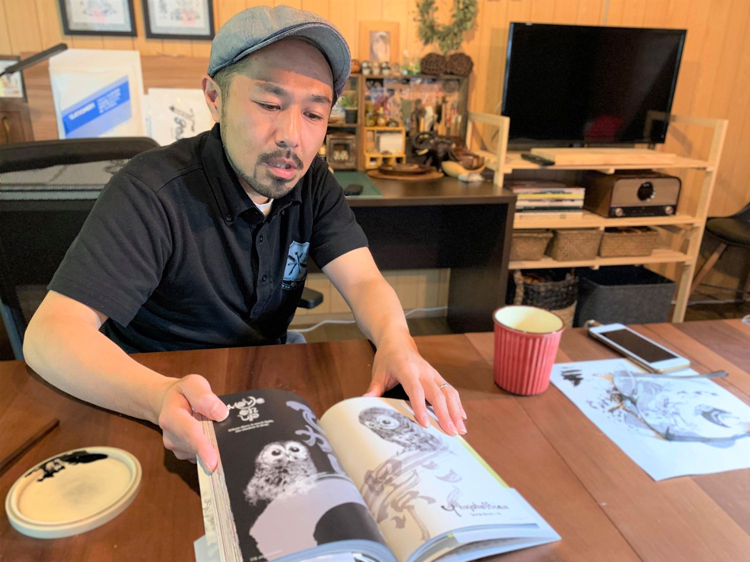 男性が机で大判の画集を開き、モノクロで描かれたフクロウのページを指差しながら作品を説明している様子の写真