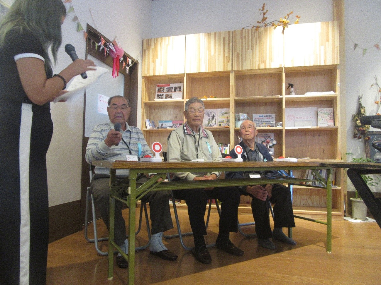 マイクを持つ高齢の男性がクイズの答えを発表しており、隣の男性二人も前を見つめて集中している様子を女性スタッフが進行しているクイズ中の写真