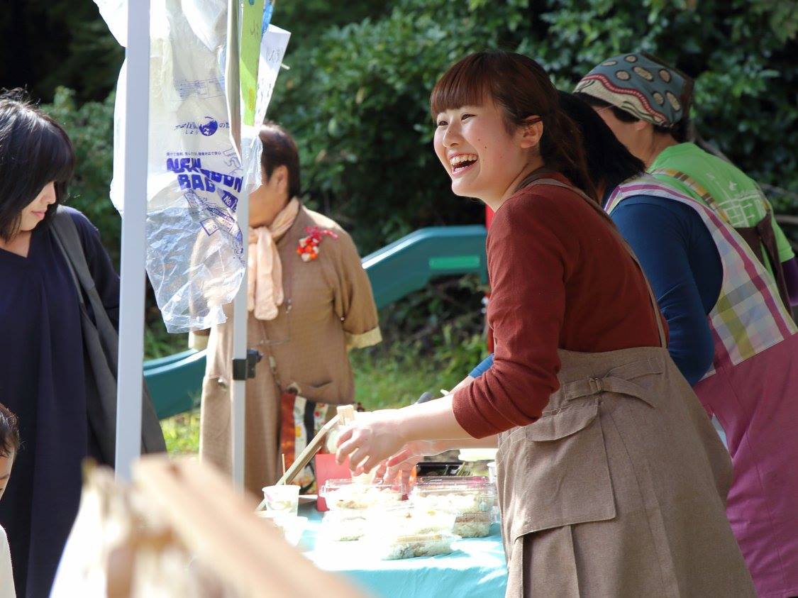 テーブルには食べ物が並んでいる屋台の前で、エプロン姿の女性が明るく笑いながら来場者に話しかけている写真