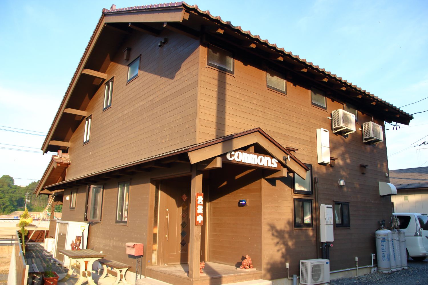 建物の周囲にエアコンの室外機やプロパンガスボンベ、自転車が置かれ、正面に「COMMONS」の白い看板が掲げられた茶色の木造二階建ての建物外観の写真