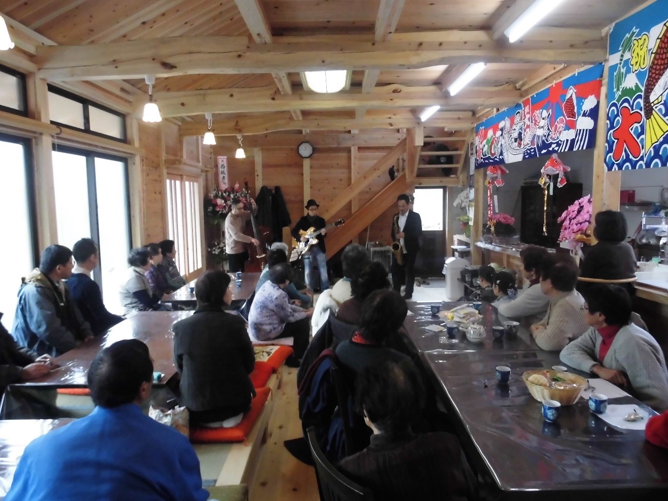 木造の室内で、長テーブルに座っている多くの人々が見守る中、JAZZ FOR TOHOKUのメンバーによるミニライブが行われている写真