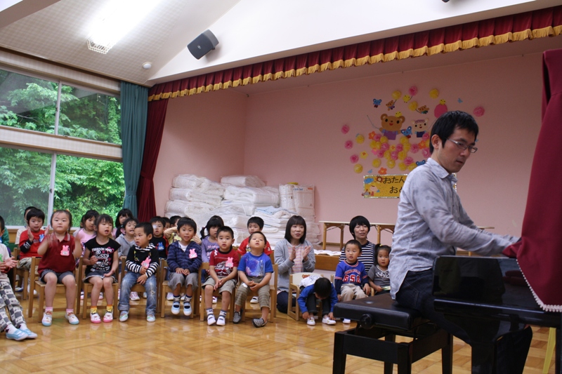 保育室で谷口博章さんがピアノを演奏し、園児たちと保育士が楽しそうに音楽を聳いている様子を写した写真