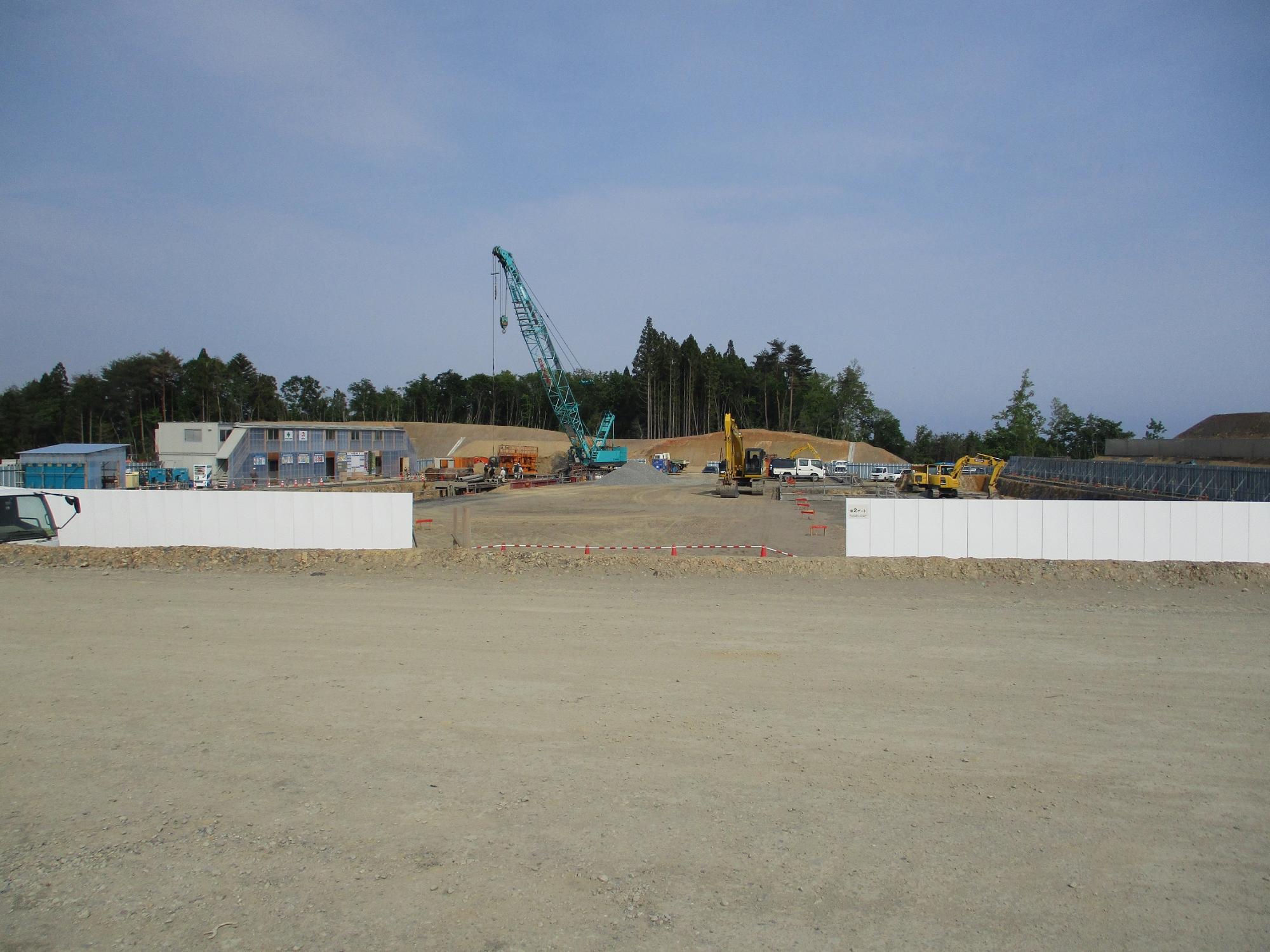 広い造成地の中央にクレーンや重機が配置されている建設現場を示しており、背景には木々が見える写真