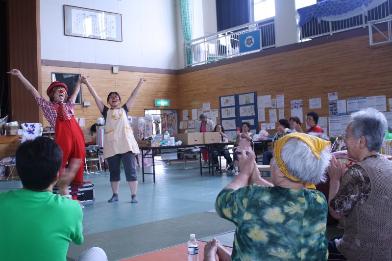 体育館の会場で、カラフルな衣装を着た二人のパフォーマーが笑顔で手を挙げて演技をし、観客の皆さんが拍手をして楽しんでいる様子を写した写真