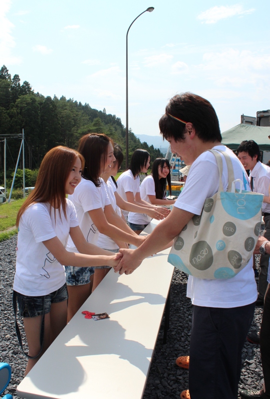 屋外の会場で、白いTシャツを着たAKB48の皆さんが長いテーブル越しに訪れた人たちと握手を交わしている様子を写した握手会の写真