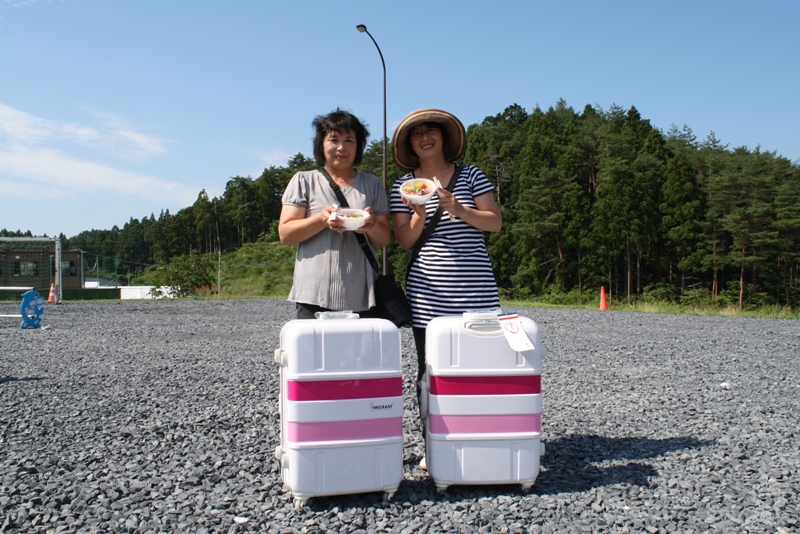 青空の下、広場で白地に濃いピンクとピンク色のラインが入ったおそろいのスーツケースの前で、二人の女性がどんぶりを手に笑顔で写っている記念写真