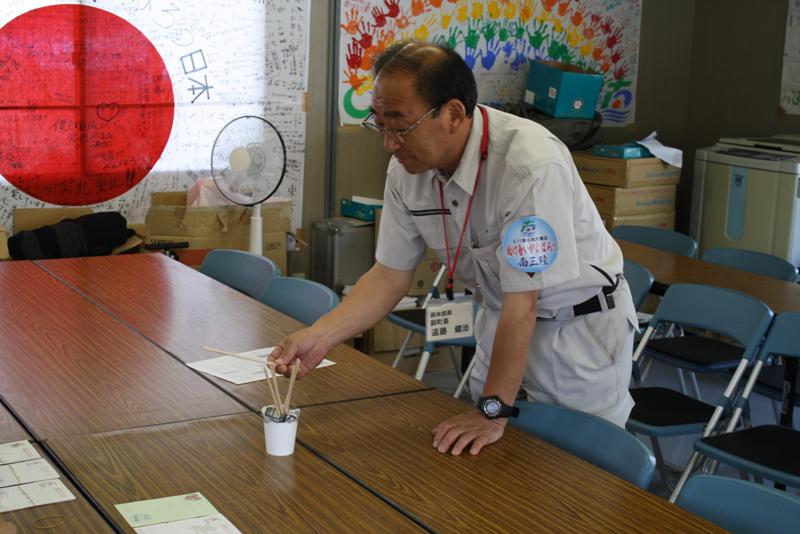 会議室の机の上に数本の棒が入った紙コップのくじが置かれ、作業着姿の男性がそのくじを1本引いている抽選会の様子を写した写真