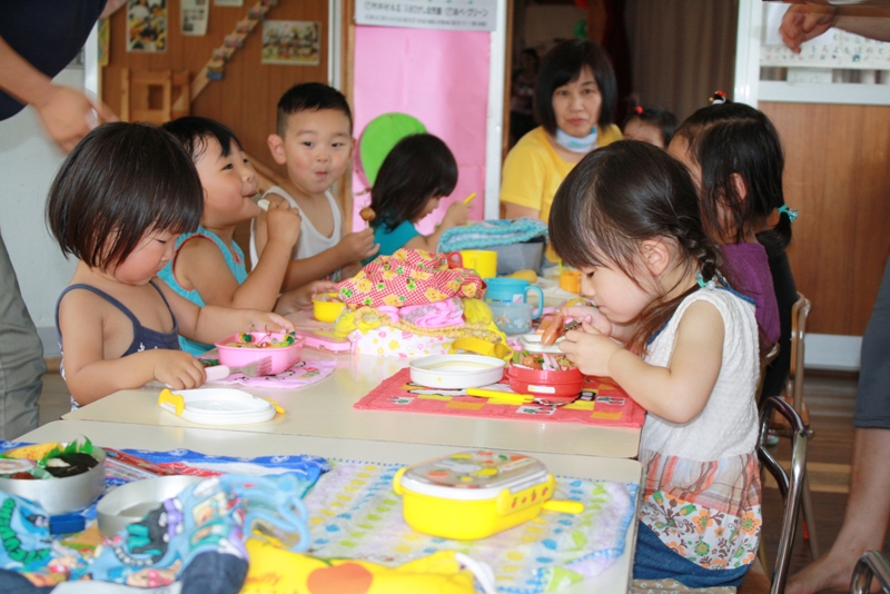 保育所の教室で、幼い園児たちがテーブルを囲んで座り、お弁当を広げて昼食を食べている様子を写した写真