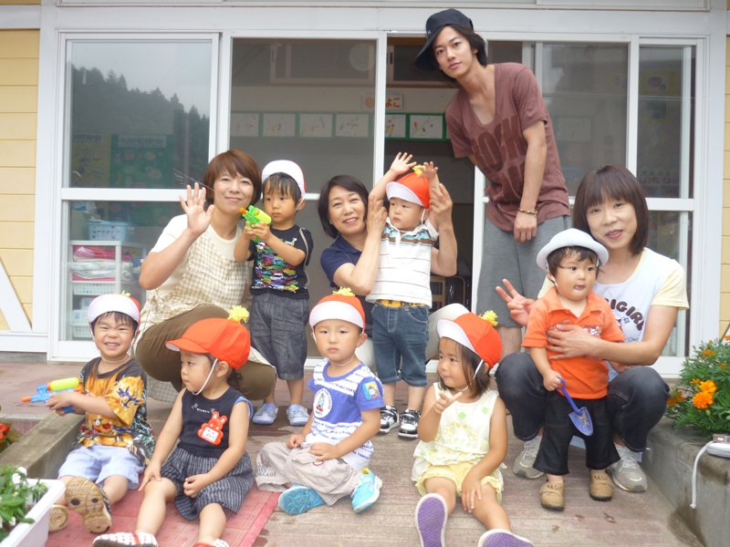 伊里前保育所の園舎前で、園児の皆さんと保育士の皆さん、佐藤健さんが一緒に笑顔で並んで写っている記念写真