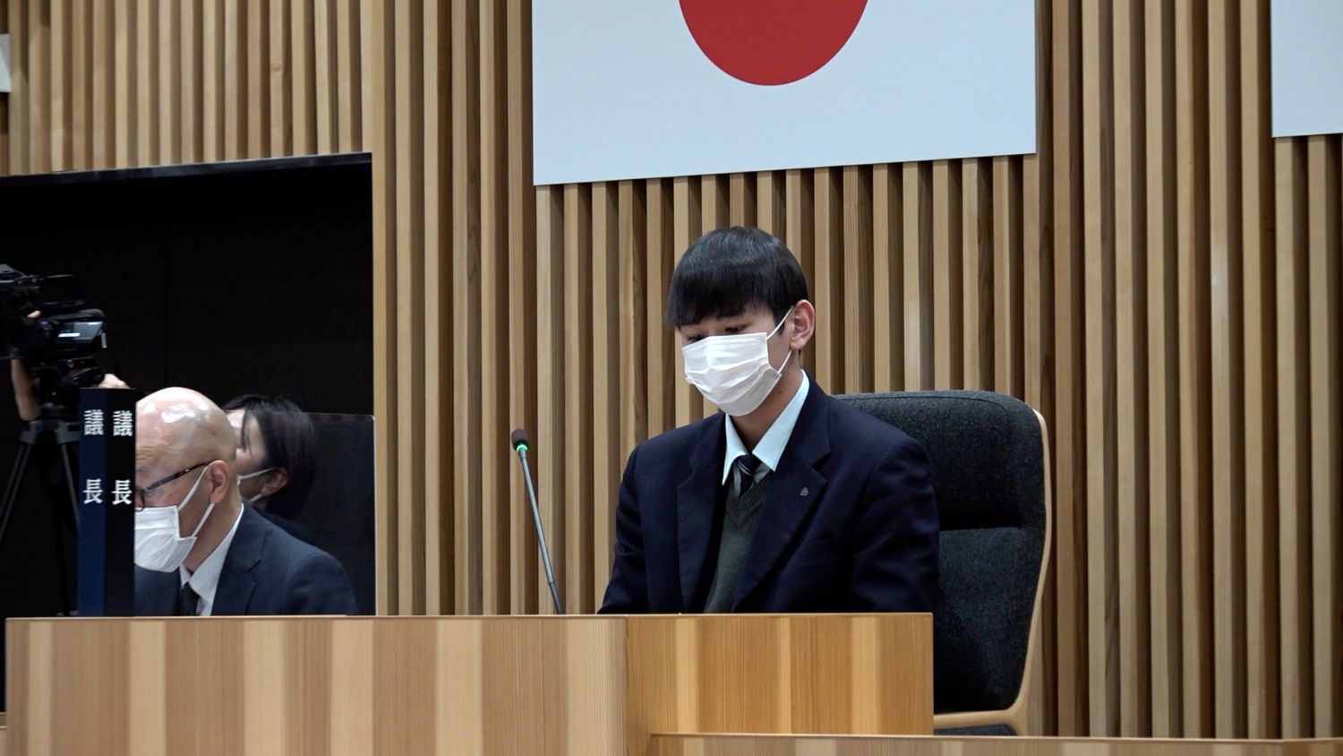 日本の国旗が掲げられた議場の壇上で、制服姿の男子学生がマスクをつけて真剣な表情で座り、左隣にはスーツ姿の大人とカメラを構える人の姿も見える写真