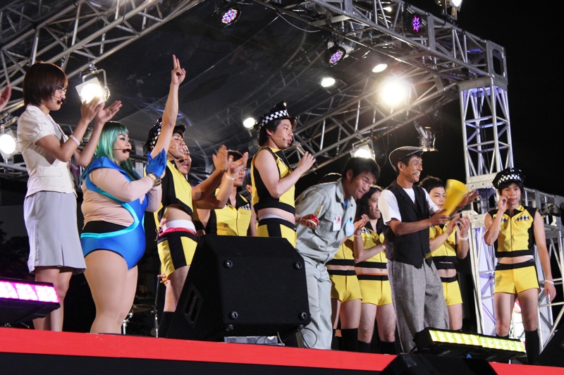 照明に照らされた特設ステージで、明石家さんまさんや黄色と黒の揃いの衣装を着た今田耕治さんをはじめ、大勢の人気芸人さんがステージライブで盛り上がっている様子を写した写真