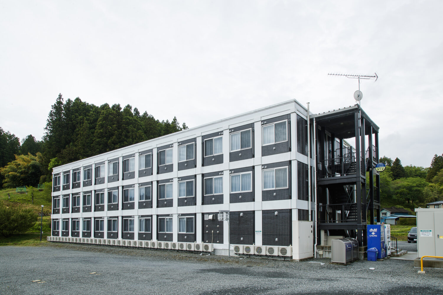 周囲には濃い緑の木々が生い茂る山が広がり、砂利の敷かれた広場の前に建っている3階建ての黒と白の外壁の学生寮の写真