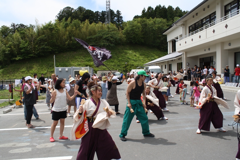 屋外の広場で、揃いの袴の衣装を着たメンバーと地域の方々が一緒によさこいを踊りを楽しんでいる様子の写真