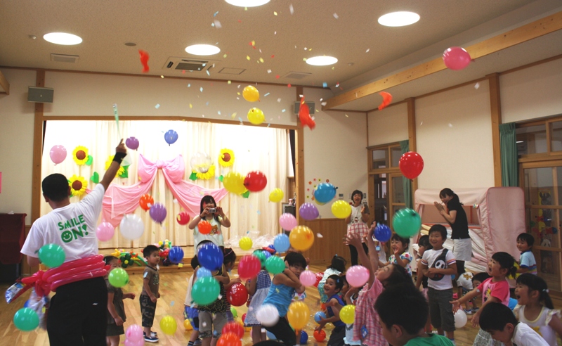 室内のホールで、『SMILE ON!』のシャツを着た梶川洋平さんがカラフルな風船と紙吹雪を降らせる中、たくさんの幼い子どもたちが手を伸ばして楽しそうに遊んでいる様子を捉えた写真