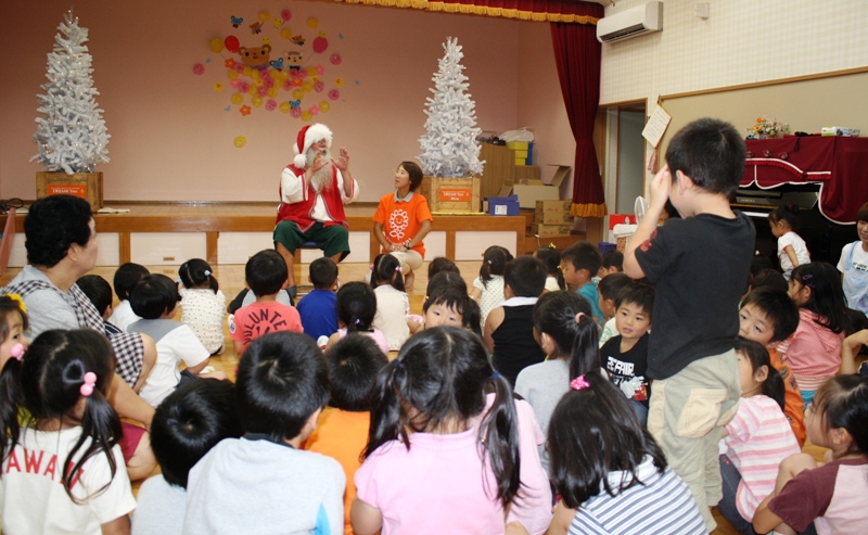 伊里前保育所のクリスマスツリーが飾られたステージで、赤い服と白いひげのフィンランド共和国のサンタクロースが座って話をし、周りにはたくさんの子どもたちが集まって真剣に話を聞いている様子を写した写真