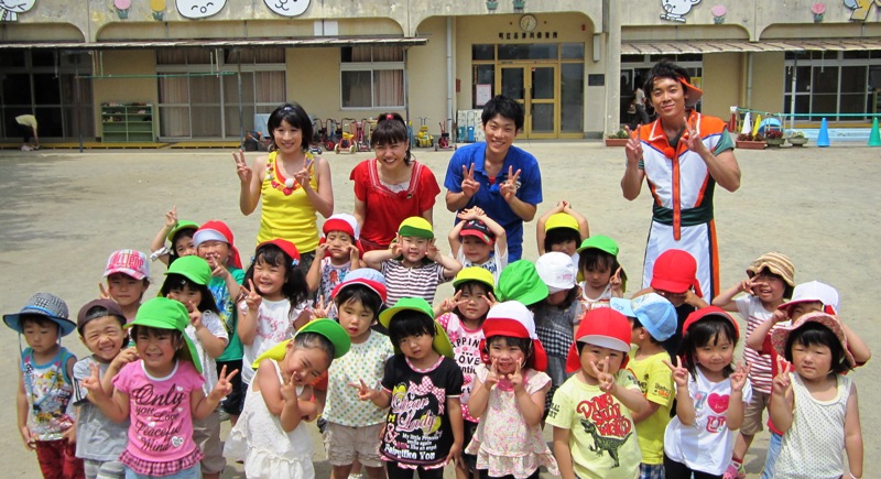園庭に子どもたちと『おかあさんといっしょ』の歌のお兄さんやお姉さん、体操のお兄さんやお姉さんが集まり、みんなで笑顔でピースサインをして写っている集合写真