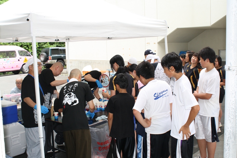 屋外に設置された白いテントの下で、黒いシャツを着た瑞巌寺の住職や関係者の方々がかき氷を作り、並んだ子どもや若者たちに渡している様子を写したかき氷の無料提供の写真