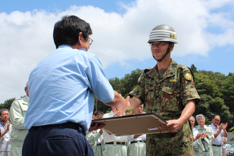 屋外で町長が自衛隊の制服を着た隊員に賞状のような額を手渡し、二人が握手を交わし、周りの人々が拍手をしている撤収セレモニーの様子を写した写真