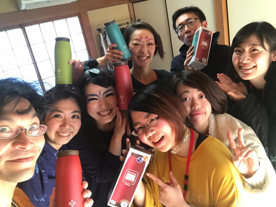 室内で8人の男女が色違いのお揃いの水筒を手に持ち、満面の笑顔でカメラに向かってポーズを取っている写真