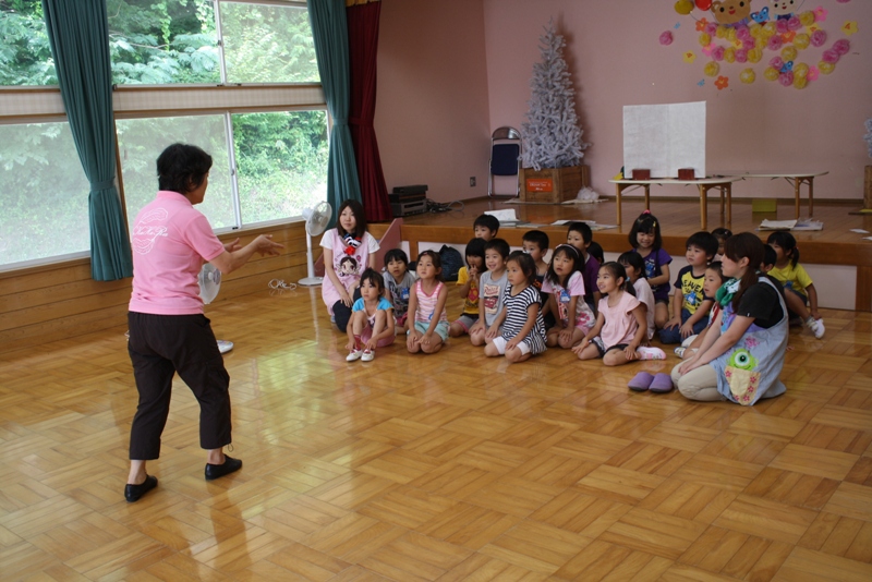 ピンク色の服を着た女性が前に立って話をし、床に座ったたくさんの子どもたちと保育士たちがその話を真剣に聞いている様子を写した写真