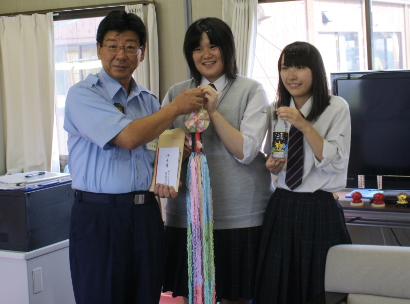 制服を着た二人の女子学生と町長が並び、色とりどりの千羽鶴の束を持ちながら笑顔で写真に収まり、男性が封筒を、女子学生の一人が仙臺（せんだい）サイダーを手にしている記念写真