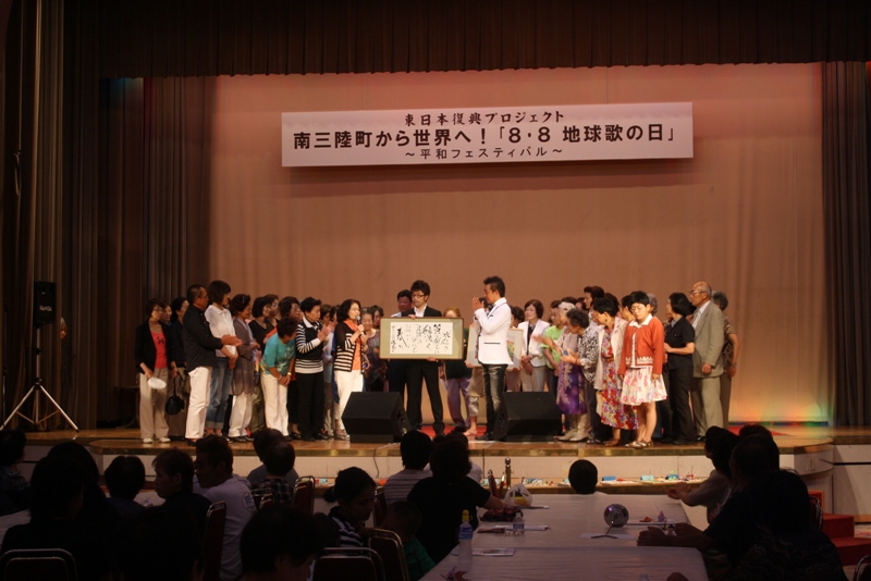 「南三陸町から世界へ！『8・8 地球歌の日』〜平和フェスティバル〜」と書かれた横断幕が掛かけられた舞台に幅広い年齢層の参加者が並び、中央で額を掲げる人物とマイクを持つ司会者が話している平和フェスティバルのの様子を写した写真