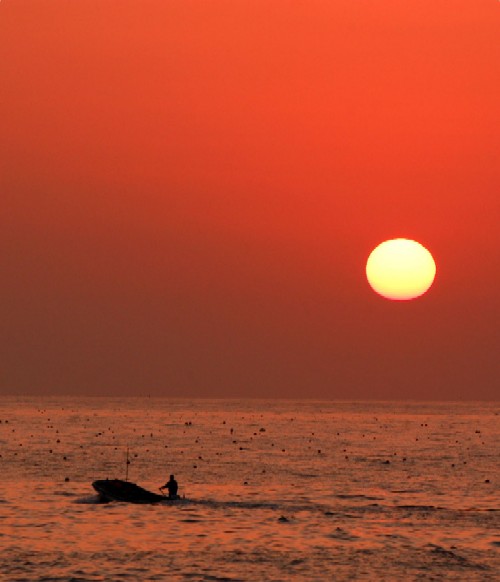 大きな太陽が、空と海を強烈な赤とオレンジ色に染め上げる中、手前の海面を小さな漁船が一人の人影を乗せて進む、ドラマチックな日の出の写真