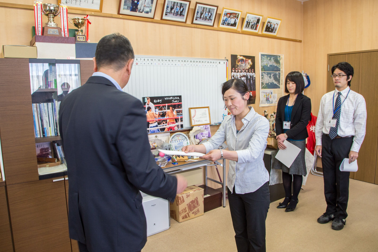 町長室で、スーツを着た男性から、女性が委嘱状を受け取っており、後ろには女性と男性が控えている写真