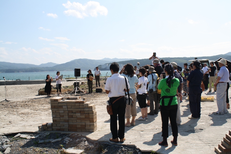 海辺で、山を背景に多くの人が集まり、スピーカーやマイクが設置された場所で撮影が行われている様子の写真