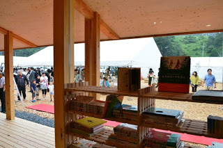 絵本や書籍が並ぶ木製の本棚越しに、屋外のテントや見学者たちの様子が見えるイベントの様子の写真