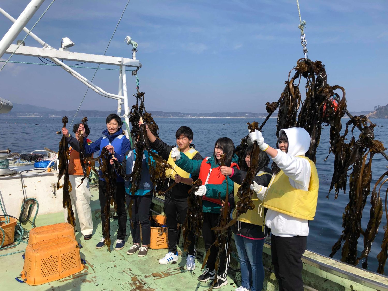 海の上の船に乗って、ジャージやライフジャケット姿の生徒たちが海藻を持ち上げて笑顔を見せている写真
