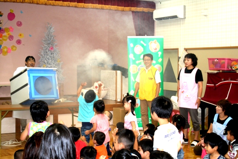 びっくり科学実験ショーで、煙が出る大きな箱から円形の煙が勢いよく飛び出し、子どもたちが前に集まって手を伸ばしながら楽しそうに反応している様子の写真
