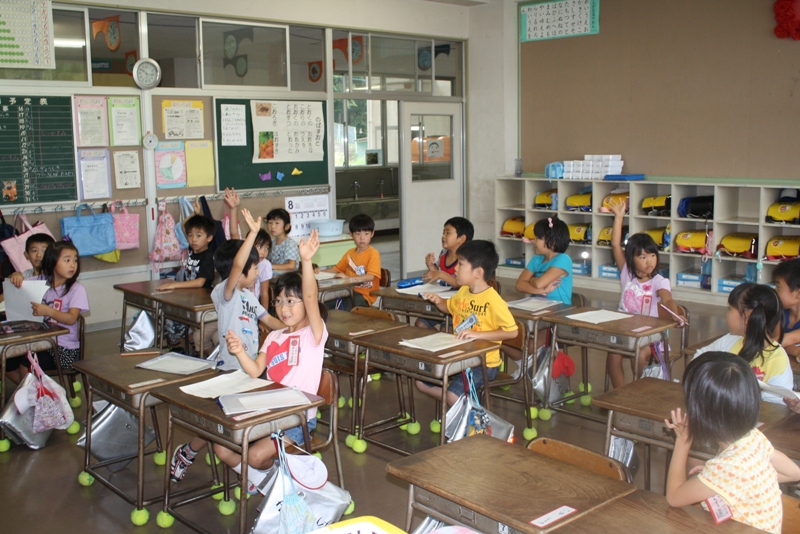 教室で席についている児童たちが先生の問いかけに元気に手を挙げて答えている様子の写真