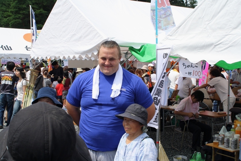 屋台やテントが並ぶイベント会場で、多くの人が集まる中、青いシャツを着た大関「把瑠都」さんが来場者と写真撮影をしている様子の写真