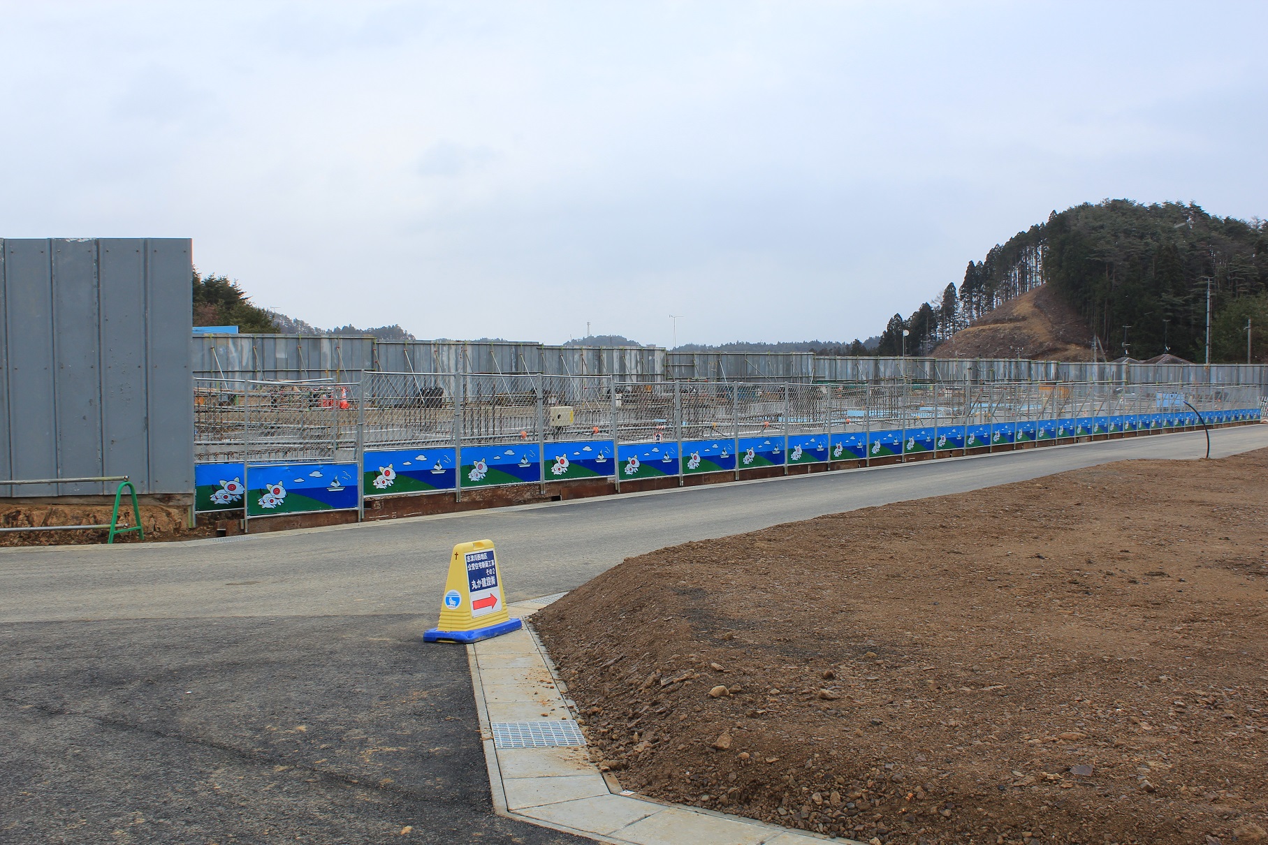 山を背景にした広い建設現場がフェンスで囲まれ、フェンスの下部には青い空と花のキャラクターが描かれた装飾が施されており、手前の道路には「車両進入禁止」の標識が設置されている写真