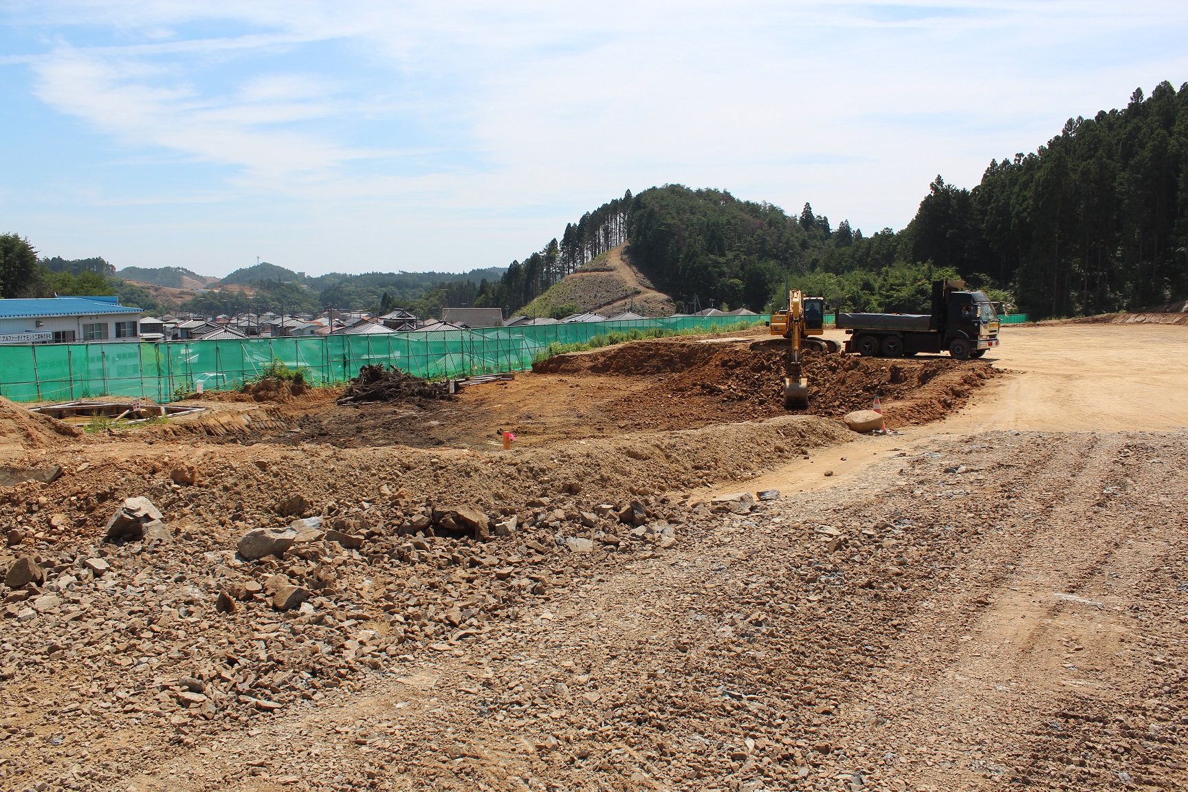 木々に囲まれた山間部の一角で造成工事が行われており、重機とダンプトラックが土を掘削する傍ら、緑のフェンスの向こうには住宅地が広がっている写真