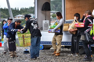 トラックの荷台の前で、黒いTシャツを着た男性が青いエプロンの人から表彰を受けており、周囲では他の参加者たちが見守る中で、表彰状のようなものを手渡している授賞式の様子の写真