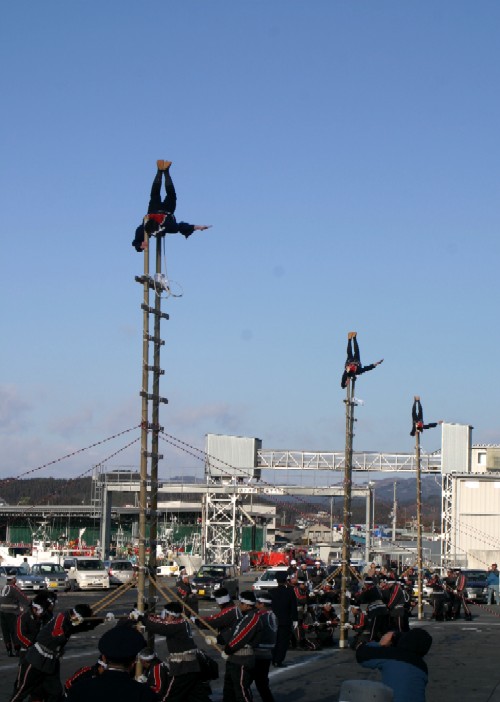 青空の下、広場のような場所で黒と赤の半被を着た人々が隊列を組み、その上空に立つ3本の長い梯子の上で1人づつ逆立ちをしてバランスをとりながら演技をしている様子の写真