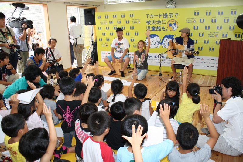 黄色い『Tカード提示で被災地に児童館を。』という横断幕の前で、土屋アンナさんを含む3人の大人がギター演奏や手振りで子どもたちと歌や音楽を楽しみ、子どもたちが手を挙げて元気に参加している様子の写真