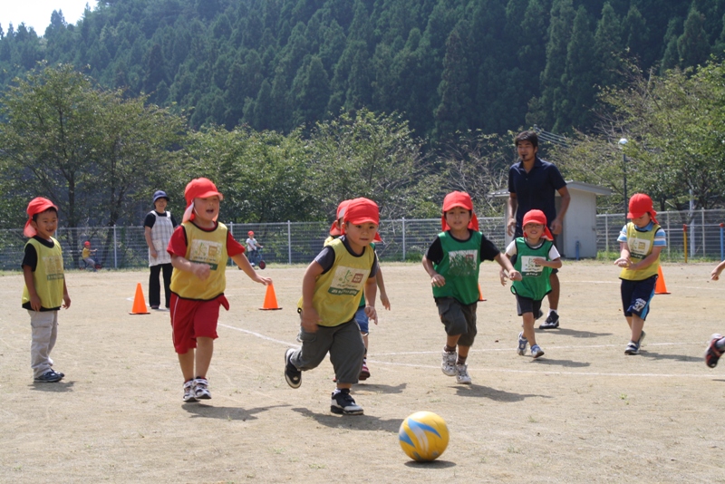 コーチの山本清治さんが見守る中、赤い帽子をかぶった子どもたちがグラウンドでボールを追いかけ、楽しそうにサッカーをしている様子の写真