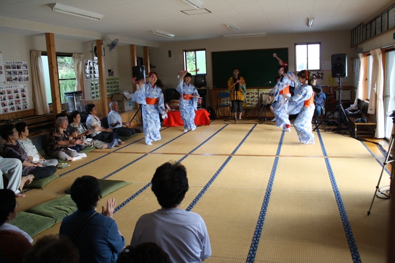 畳の部屋で、青と白の模様の浴衣にオレンジ色の帯を締めた四名の踊り手が民謡の踊りを披屋しており、周囲で座って見ている観客が手拍子をしながら楽しんでいる民謡キャラバンの様子の写真