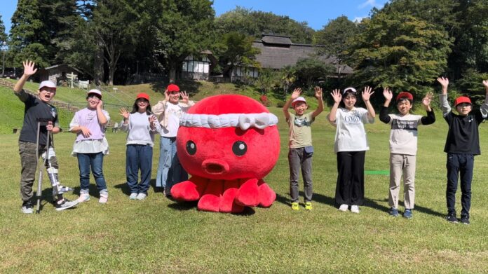 赤いたこ型のマスコットキャラクターを中心に、外で整列して笑顔で手を振っている子どもたちが青空の下で撮影されている写真